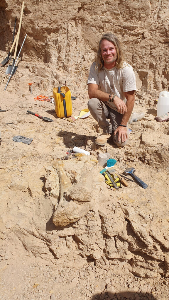 Dr Phil Bell working in the field. Smiling at the camera while kneeling on the ground. He is surrounded by his tools and equipment. 