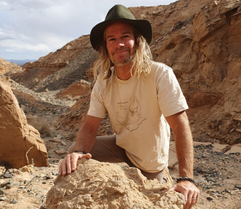 Dr Phil Bell - University of New England’s Palaeontologist and Associate Professor. He is kneeling on the ground in the field leaning on a rock with two hands.