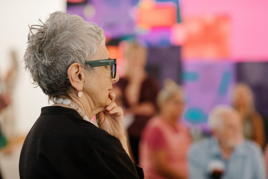 A person with short gray hair stands in the foreground of an art gallery, wearing a beaded necklace and holding a hand near their chin as if observing something thoughtfully. In the background, several people sit or stand while colorful abstract artworks hang on the wall.